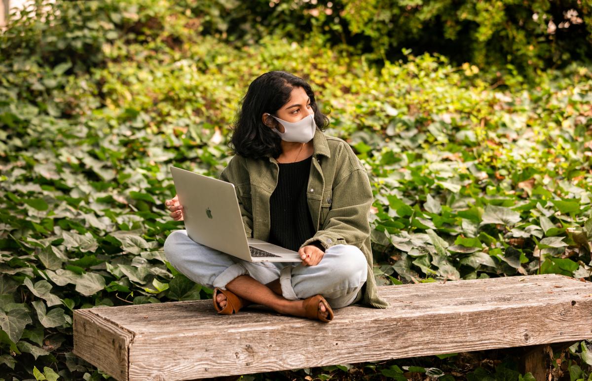 A student, wearing a mask, sits on her laptop and looks off into the distance